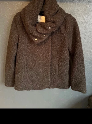 Brun teddyjacka från Jacqueline de Yong - Mysig svart  teddyjacka från Jacqueline de Yong med stor krage och knappar framtill. Jackan har två fickor och är tillverkad i ett fluffigt teddy-material som håller dig varm under kyliga dagar. Perfekt för dig som gillar en avslappnad men trendig stil.