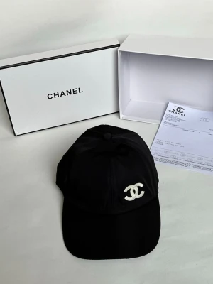 Svart keps från Chanel med logga - Snygg svart keps från Chanel med den ikoniska vita CC-loggan broderad framtill. Klassisk böjd skärm och justerbar rem bak. Tillverkad i ett slätt tyg som ger en exklusiv känsla. Perfekt accessoar för att lyfta din outfit.