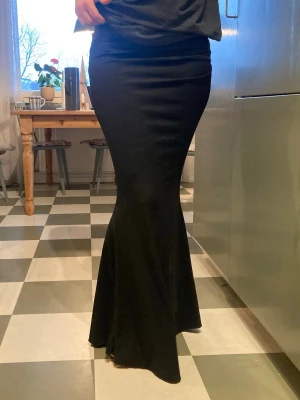 Svart lång tight kjol med utsvängd nederdel - Uutsvängd nederdel som ger en snygg siluett. Kjolen är enkel och stilren, perfekt att matcha med olika toppar. Jättebra skick, som ger en fantastisk siluett och är som den lovar - läng. Jag är 173cm och den går att ha på både kortare och längre. Den har lite scrunch på vänstra höften som gör så kroppen får en otrolig form. Superskönt material som inte är sånt sportmaterial. Jag önskar den verkligen ett varmt nytt hem, som jag inte längre kan erbjuda. Priset är diskuterbart.