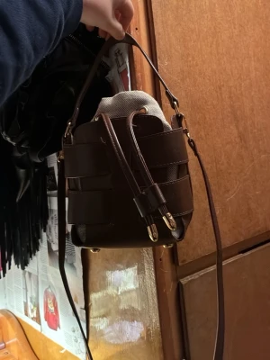 Brun bucket handväska i skinn - Snygg brun bucket handväska med flätad design och guldfärgade detaljer. Väskan har både handtag och axelrem samt en tygpåse inuti som stängs med snörning. Perfekt för dig som vill ha en unik och trendig accessoar.