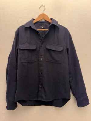 Mörkblå overshirt med fickor - Stilren mörkblå overshirt i storlek M från H&M med två bröstfickor och knappar framtill. Skön och enkel att matcha, perfekt lager på lager. Skriv om ni har några fler frågor!