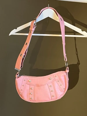 Rosa axelväska med nitar - Trendig rosa axelväska i syntetmaterial med croco-mönster och silverfärgade nitar. Väskan har justerbart axelband med metallspännen och detaljer som ger en cool vibe. Perfekt accessoar för att lyfta din outfit. Helt oanvänd, köpt från ASOS🩷