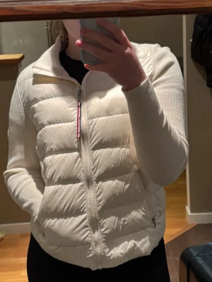 Vit quiltad jacka Tommy Hilfiger - Snygg beige quiltad jacka från Tommy Hilfiger med ribbade stickade ärmar och krage. Jackan har dragkedja framtill, två fickor med dragkedja och ett diskret logomärke på ärmen. Perfekt för lager på lager under höst och vår.