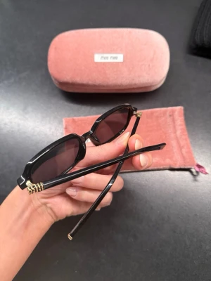 Svarta solglasögon från Miu Miu - Snygga svarta solglasögon från Miu Miu med rektangulära bågar och mörka linser. Detaljer i guld vid gångjärnen ger en lyxig touch. Kommer med rosa fodral och matchande tygpåse. Perfekta för dig som vill ha en trendig accessoar. Kvitto finns ej då det är en present. Använda fåtals gånger 