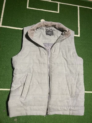 Ljusgrå dunväst från Dressmann - Stilren ljusgrå dunväst från Dressmann med dragkedja framtill och två snedställda fickor. Kragen har ett snyggt manchestertyg på insidan som ger extra detalj. Perfekt för lager på lager och enkel att matcha med olika outfits.