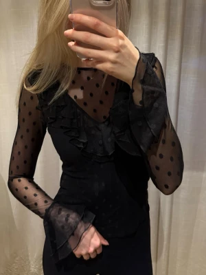 Svart meshblus med volang och prickar - Snygg svart meshblus med långa ärmar, volangdetaljer och prickigt mönster. Transparenta ärmar och volanger vid ärmslut och halsringning. Har ett linne under på bilderna. Se sista bilden för att se exakt hur den ser ut.