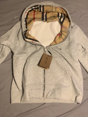 Burberry hoodie zip up (billigt) - Snygg grå zip hoodie från Burberry med klassiskt beige, rött och svart rutmönster i huvan och insidan. Hoodien har dragkedja framtill, två fickor och ribbade muddar. Perfekt för dig som vill ha en stilren men ändå lyxig look. Anledningen varför jag säljer den är för den va för stor för mig helt ny direkt från paketet bästa pris. (Har endast provat en gång) pris kan diskuteras. Välj instabox för snabb leverans