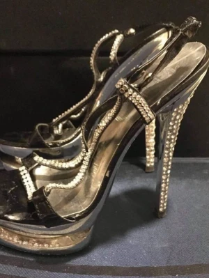 Svarta pumps med strass och hög klack - Glammiga svarta pumps med hög klack och plattform, dekorerade med glittrande strass på remmar och klack. Skorna har en spetsig siluett och öppen tå, perfekta för att sticka ut. Materialet är syntet med lackad finish.passar st /38-39