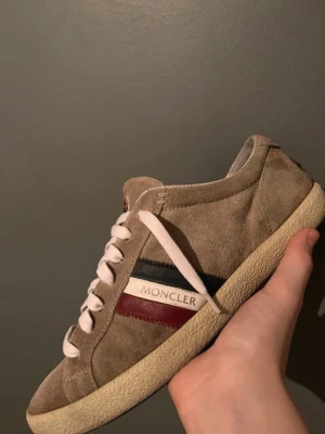 Beige Moncler sneakers i mocka - Snygga beige sneakers från Moncler i mjuk mocka med vita snören och rund tå. Sidan har Monclers logga och klassiska ränder i rött och blått. Skorna har platt sula och en stilren, tidlös design som passar till det mesta. Det är en liten fläck på sulan.