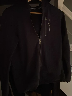 Svart hoodie från Sail Racing - Säljer en svart hoodie från Sail Racing med dragkedja framtill och logga på bröstet. Tröjan har huva och ribbade muddar vid ärmslut och nederkant. Perfekt för dig som gillar stilrena och sportiga plagg. Jag köpte den på Kid Brand store