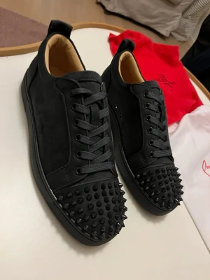 Svarta sneakers från Christian Louboutin - Säljer ett par gråa sneakers från Christian Louboutin med mattsvarta nitar på tån och klassisk röd sula. Skorna har snörning och är tillverkade i mocka och skinn för en lyxig känsla. Perfekta för dig som vill sticka ut med en edgy stil.
