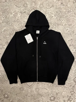 Svart zip hoodie från Arte - Snygg svart hoodie från Arte med dragkedja framtill och broderad logga på bröstet. Klassisk huva och känguruficka gör den både stilren och praktisk. Tillverkad i mjukt material som känns skönt mot huden. Perfekt för en avslappnad streetwear-look.