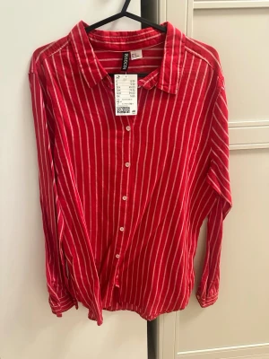 Rödvit randig skjorta från H&M - Snygg röd skjorta från H&M med vita vertikala ränder. Skjortan har klassisk krage, knäppning framtill och långa ärmar. Tillverkad i mjuk bomull som känns skön mot huden. Perfekt för dig som vill ha en färgstark och trendig look.