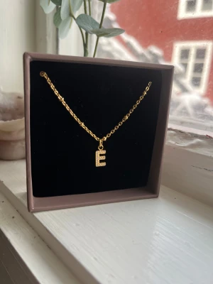 Guldhalsband med bokstav E - Snyggt halsband i guldton med en kedja och ett hängsmycke i form av bokstaven E, dekorerad med små gnistrande stenar. Perfekt accessoar för dig som vill ha något personligt och trendigt. Säljer för jag inte andvänder guld, aldrig andvänd, fått den i present i missförstånd att jag inte gillar guld. 