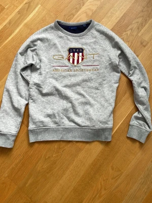 Grå sweatshirt från Gant - Säljer en grå sweatshirt från Gant med broderad logga och sköld på bröstet. Tröjan har rund halsringning, ribbade muddar och texten 'American Sportswear' framtill. Perfekt för en avslappnad och sportig stil.