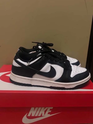 Nike Dunk Low svart/vit sneakers - Nike Dunk Low sneakers i svart och vitt med klassisk rund tå och snörning. Skorna har en platt sula och är tillverkade i skinn och syntet. Ikonisk Swoosh-logga på sidan och perforerad tåbox för extra ventilation. Perfekt för dig som gillar streetwear och stilrena färger.
