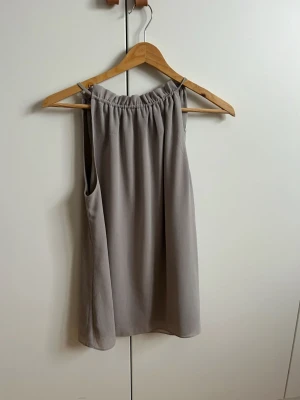 Grå ärmlös topp/blus - Stilren grå linnetopp med rak passform som man knyter bak. Toppen är ärmlös och har en enkel, clean look som passar perfekt till många olika outfits. Tillverkad i ett lätt och luftigt material.