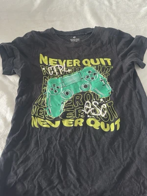 Svart gamer t-shirt med tryck H&M - Cool svart t-shirt från H&M med stort färgglatt tryck av en grön spelkontroll och texten 'NEVER QUIT' i gult och vitt. Perfekt för dig som älskar gaming och vill sticka ut med en unik design. Klassisk passform och rund hals.