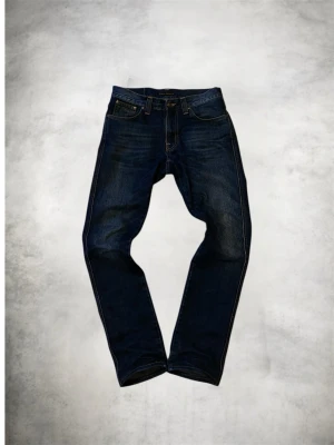 Mörkblå Nudie Jeans Slim Fit - Snygga mörkblå jeans från Nudie Jeans med klassisk femficksdesign och kontrastsömmar. Modellen har en slim passform och raka ben. Tillverkade i slitstarkt bomullsmaterial med subtila slitningar för en cool look. Strlk 31-32/34. (Uppsydda ifrån 36) 