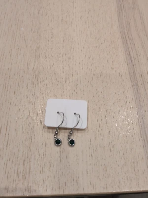 Silverörhängen med grön sten i rostfritt stål  - Snygga örhängen i silverfärg med rund grön sten som hänger i en enkel krok. Perfekta för att ge en färgklick till din outfit. Materialet ser ut att vara metall och stenen har en glittrande finish.