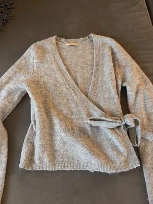 Grå omlottkofta från Gina Tricot - Mysig grå stickad omlottkofta från Gina Tricot med knytband i midjan och djup v-ringning. Koftan har långa ärmar och är tillverkad i ett mjukt, fluffigt material som känns skönt mot huden. Perfekt att slänga på sig när det är lite kyligt ute.