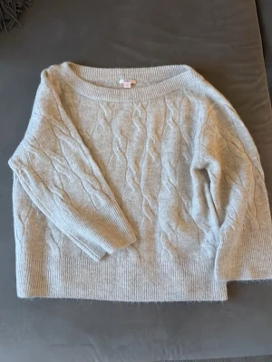 Ljusgrå kabelstickad tröja Gina Tricot - Mysig ljusgrå kabelstickad tröja från Gina Tricot med bred ribbad kant vid hals, ärmslut och nederkant. Tröjan har en avslappnad passform och är perfekt för kyliga dagar. Klassisk design med diskret mönster i garnet. 