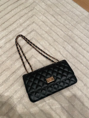 Svart quiltad axelväska med guldkedja - Säljer en svart quiltad handväska med guldfärgad vridknäppe och kedjerem med inslag av svart läder. Klassisk rektangulär form och elegant design som passar till många olika looks. Perfekt för dig som gillar tidlösa accessoarer.