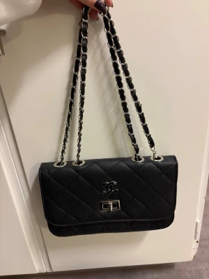 Svart väska - Helt ny, svart väska med ställbart axelband. Så man kan ha den både som hand-och axelväska. Silverdetaljer. Helt ny, men skada nere i botten på insidan av väskan (se sista bilden). Går säkert att limma om man vill, men inget som syns eller störs, eftersom det är på insidan. Även ett mindre fack på insidan. Inte en skråma i övrigt. Bara stått bland mina andra väskor. Mått ca 25cm lång och ca 16cm hög.