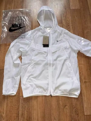 Vit vindjacka med huva från Nike React - Snygg vit vindjacka från Nike React med huva och dragkedja framtill. Jackan har diskret Nike-logga på bröstet, två sidofickor och ventilerande meshpanel på ryggen. Tillverkad i lätt polyester, perfekt för blåsiga dagar. Enkel och stilren design.