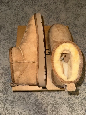 Beige UGG boots i mocka med ullfoder - De populära beige UGG skorna i mjuk mocka med varmt foder i fårskinn. Helt oanvända, säljer pga fel storlek. Ultra mini modell.