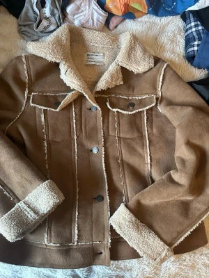 Brun teddyjacka från Brownie - Snygg brun teddyjacka från Brownie med krage och detaljer i beige teddyfoder. Jackan har knappar framtill, två bröstfickor och uppvikta ärmslut med mjukt foder. Perfekt för dig som vill ha en mysig och trendig look i höst och vinter.