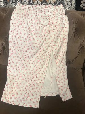 Vit blommig långkjol med snörning - Söt vit långkjol med små rosa blommor och snörning framtill som ger en snygg rynkad effekt. Kjolen har en slits framtill och är perfekt för dig som gillar romantisk och feminin stil. ( finns topp om man vill köpa med till den) 