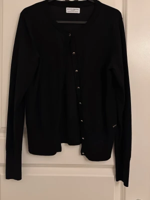 Svart kofta från Holly & White - Stilren svart kofta från Holly & White med rund halsringning och knäppning framtill. Långärmad modell i mjukt, tunt material som passar perfekt till lager-på-lager. Klassisk och enkel design som funkar till det mesta.