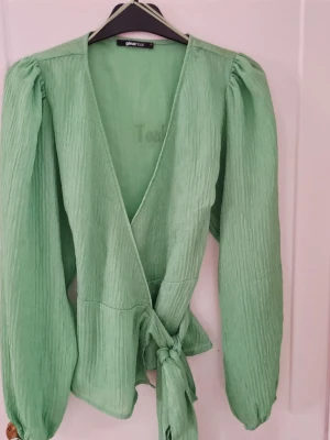 Grön omlottblus från Gina Tricot - Snygg ljusgrön omlottblus från Gina Tricot med puffiga långärmar och knyt i midjan. Blusen har v-ringning och är gjord i ett lätt, ribbat material som ger en avslappnad men trendig look.