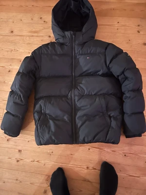 Svart pufferjacka från Tommy Hilfiger - Svart pufferjacka från Tommy Hilfiger med huva och dragkedja framtill. Jackan har diskret logga på bröstet och stora broderade bokstäver på insidan. Perfekt för kalla dagar och riktigt snygg till streetwear-stilen.