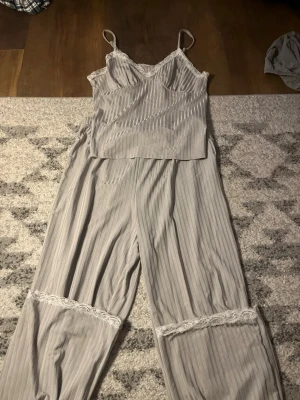 Grårandig pyjamas med spetsdetaljer - Tvådelad pyjamas i ljusgrått med tunna vita ränder. Linnet har smala axelband och spetskant vid urringningen. Byxorna är vida med spetsdetaljer vid knäna. Mjuk och luftig känsla, perfekt för chillkvällar hemma.