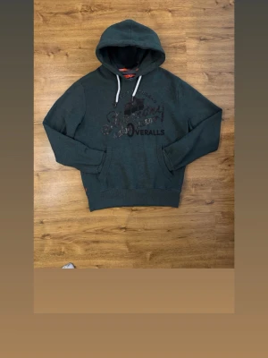 Mörkgrön hoodie från Superdry - Denna snygga mörkgröna hoodie från Superdry har ett brösttryck och vita dragsnören i huvan.  Den klassiska kängurufickan framtill och det mjuka materialet ger en skön känsla mot huden. Perfekt för en avslappnad och cool look. Den är storlek L 