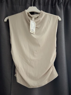 Beige ärmlös topp från H&M - Stilren beige ärmlös topp från H&M med hög hals och snygg rynkdetalj på sidan. Tillverkad i mjuk polyester som ger en skön känsla och lätt glans. Perfekt att matcha med jeans eller kjol för en trendig look.
