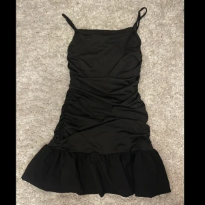 Svart kort klänning med volang - Snygg svart kort klänning med tunna axelband och volang längst ner. Klänningen har en figurnära passform och lätt rynkade detaljer på sidorna. Perfekt för dig som gillar en enkel men trendig look.