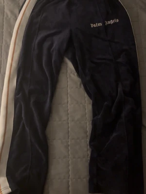 Palm Angels mörkblå mjukisbyxor - Mörkblå mjukisbyxor från Palm Angels med vit sidoremsa och beige rand. Byxorna har dragkedja vid benslut och logga broderad framtill. Tillverkade i mjuk polyester med loose passform, perfekta för en avslappnad streetwear-look.