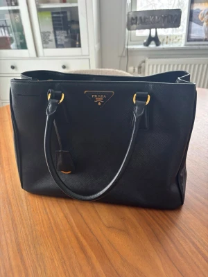 Svart handväska från Prada i skinn - Klassisk svart handväska från Prada i elegant skinn med guldfärgade metalldetaljer och dubbla handtag. Väskan har en stilren, fyrkantig form och ikonisk Prada-logga framtill. Perfekt för dig som gillar lyxiga accessoarer med tidlös design.
