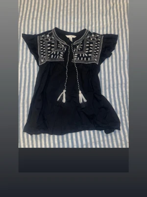 Svart blus med broderi från HM - Svart blus med vita broderade detaljer över bröstet och axlarna. Toppen har korta ärmar och dekorativ snörning med tofsar framtill. Perfekt för dig som gillar boho-stil och vill ha något unikt i garderoben.