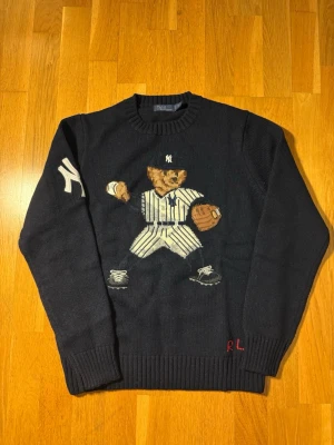 Ralph Lauren New York tröja  - Mörkblå stickad tröja från Ralph Lauren med ikonisk björn i basebolloutfit på bröstet och vit New York Yankees-logga på ärmen. Tröjan har ribbstickade muddar och RL-broderi i rött vid nederkanten. 