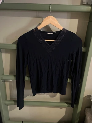 Svart långärmad topp med spets - Snygg svart långärmad topp med v-ringning dekorerad med spetsdetaljer. Toppen har en smickrande passform och är perfekt för dig som gillar stilrena plagg med en twist. Materialet känns mjukt och stretchigt, vilket gör den bekväm att bära hela dagen.