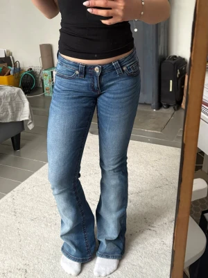 Blå bootcut jeans från Gina tricot - Säljer ett par klassiska blå jeans med låg midja och bootcut passform. Jeansen har fem fickor, knappgylf och är tillverkade i slitstarkt bomullsdenim. Perfekta för dig som gillar en tidlös och avslappnad stil.