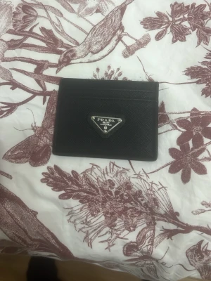 Svart kortfodral från Prada i skinn - Snyggt svart kortfodral från Prada i exklusivt skinn med metallogo framtill. Smidig och stilren design med flera kortfack, perfekt för att hålla koll på dina viktigaste kort. Klassisk och tidlös accessoar från ett lyxigt modehus.
