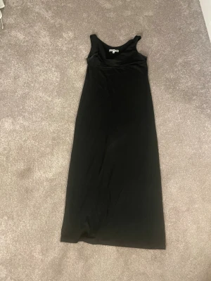 Svart långklänning i bomull - En enkel och stilren svart långklänning med ärmlös design och rund halsringning. Klänningen är gjord i mjuk bomull och har en rak passform som ger en avslappnad look. Perfekt att styla med sneakers eller sandaler. Den har lite extra tyg vid halsen så den blir lite ”hängig” på ett snyggt sätt