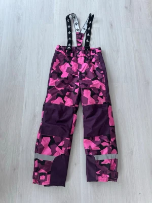  skidbyxor från Cross Sportwear - Skidbyxor från Cross Sportwear med rosa och lila mönster. Byxorna har hängslen, förstärkta knän och reflexdetaljer vid bensluten. Perfekta för vintersport och utomhusaktiviteter.
