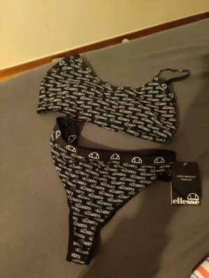 Svart thong set från Ellesse - Snyggt svart underklädesset från Ellesse med matchande topp och trosa i thong-modell. Setet har vitt logomönster över hela tyget och tunna axelband på toppen. Materialet är mjukt och stretchigt jerseytyg för skön passform.