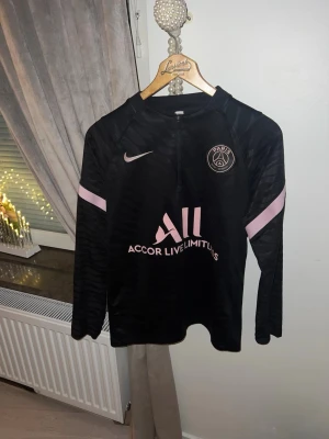 Svart PSG träningströja  - Snygg svart långärmad träningströja från PSG med rosa detaljer och halv dragkedja. Tröjan har PSG-logga och Nike Swoosh på bröstet samt sponsortryck fram och bak. Tillverkad i ett lätt och funktionellt material som passar perfekt till träning.
